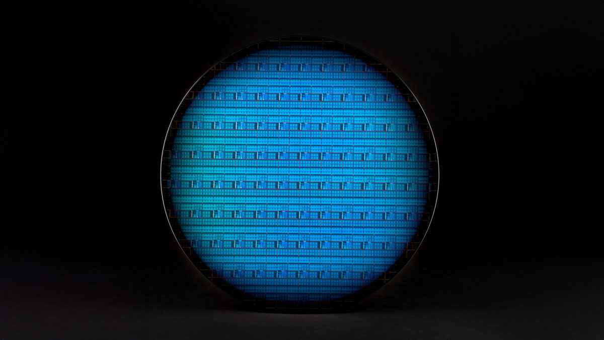 A close-up view of a silicon wafer with intricate blue circuitry patterns against a dark background. The circular wafer showcases a grid-like design, emphasizing the precision and complexity of microchip technology.