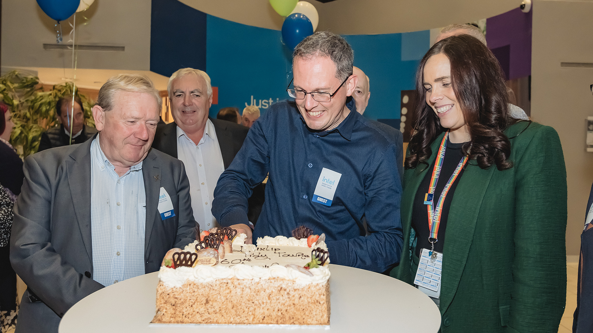 A group of people stand around a cake on a table. The man in the center, wearing glasses, slices the cake with a smile. Others watch and smile. The cake has decorative icing and the words Thank you are visible. Balloons are in the background.
