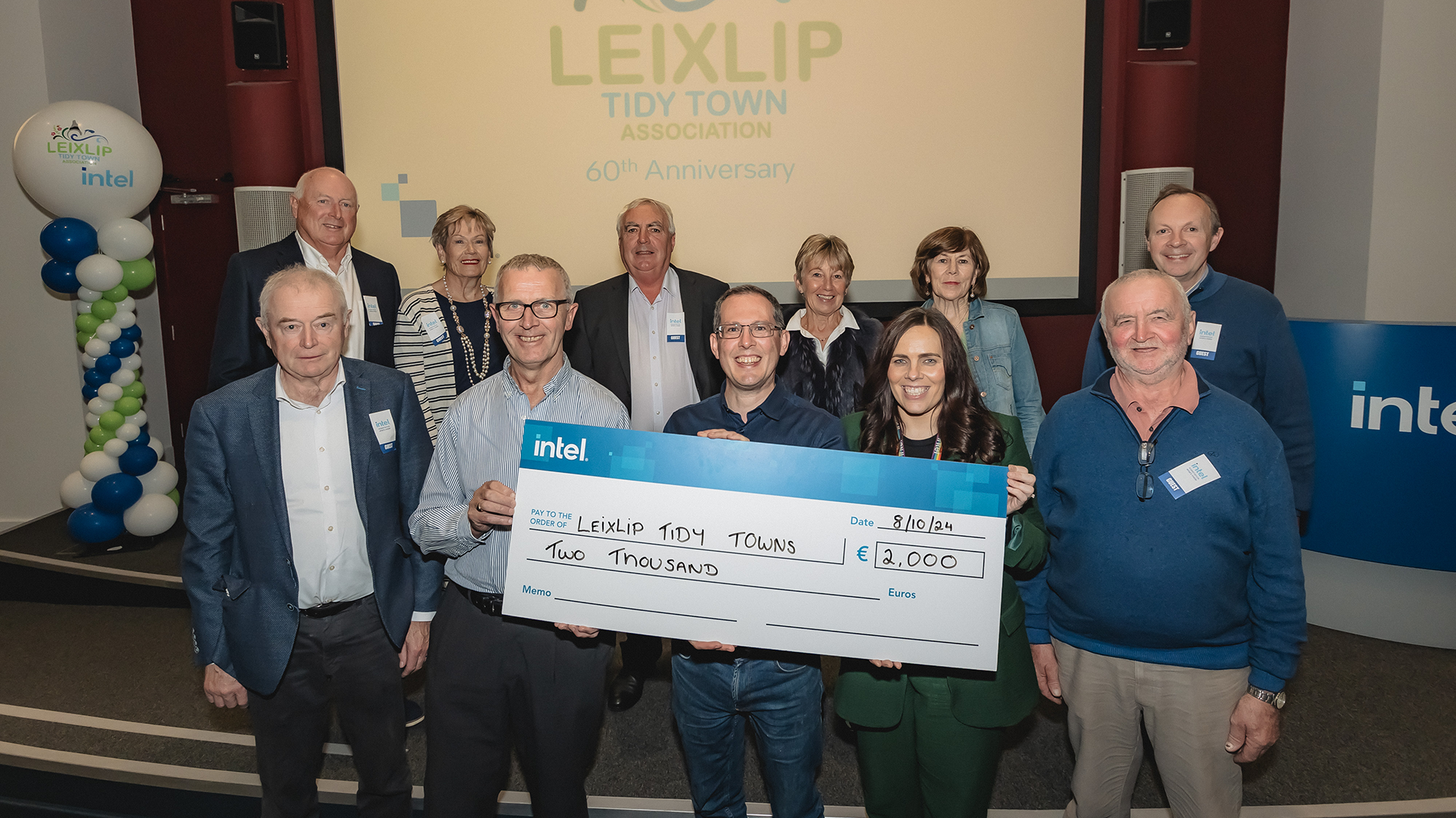 A group of eleven people stands on a stage holding a large ceremonial check for €2,000 presented to Leixlip Tidy Towns. They are smiling, and balloons are visible in the background.