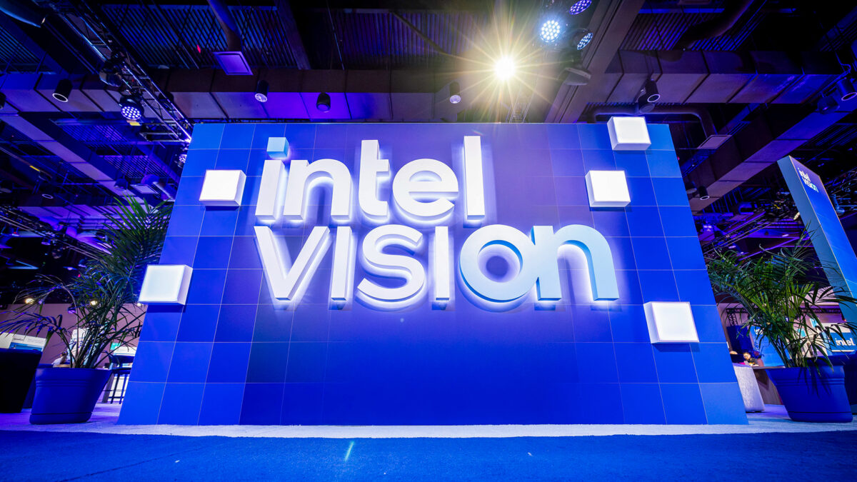 A large blue display with the words intel vision in white, illuminated letters, set against a modern, industrial-style ceiling with lighting and plants on the sides.