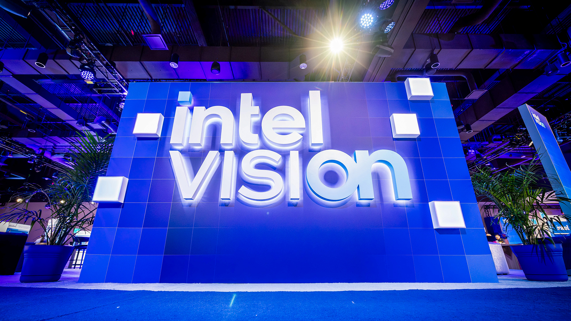 A large blue display with the words intel vision in white, illuminated letters, set against a modern, industrial-style ceiling with lighting and plants on the sides.