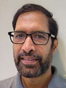 a man with short dark hair, beard and glasses wearing a polo-style shirt, looks at the camera